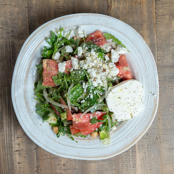 Vibrant salad with greens, tomatoes, chickpeas, crumbled feta, and a large cheese slice on a white plate.