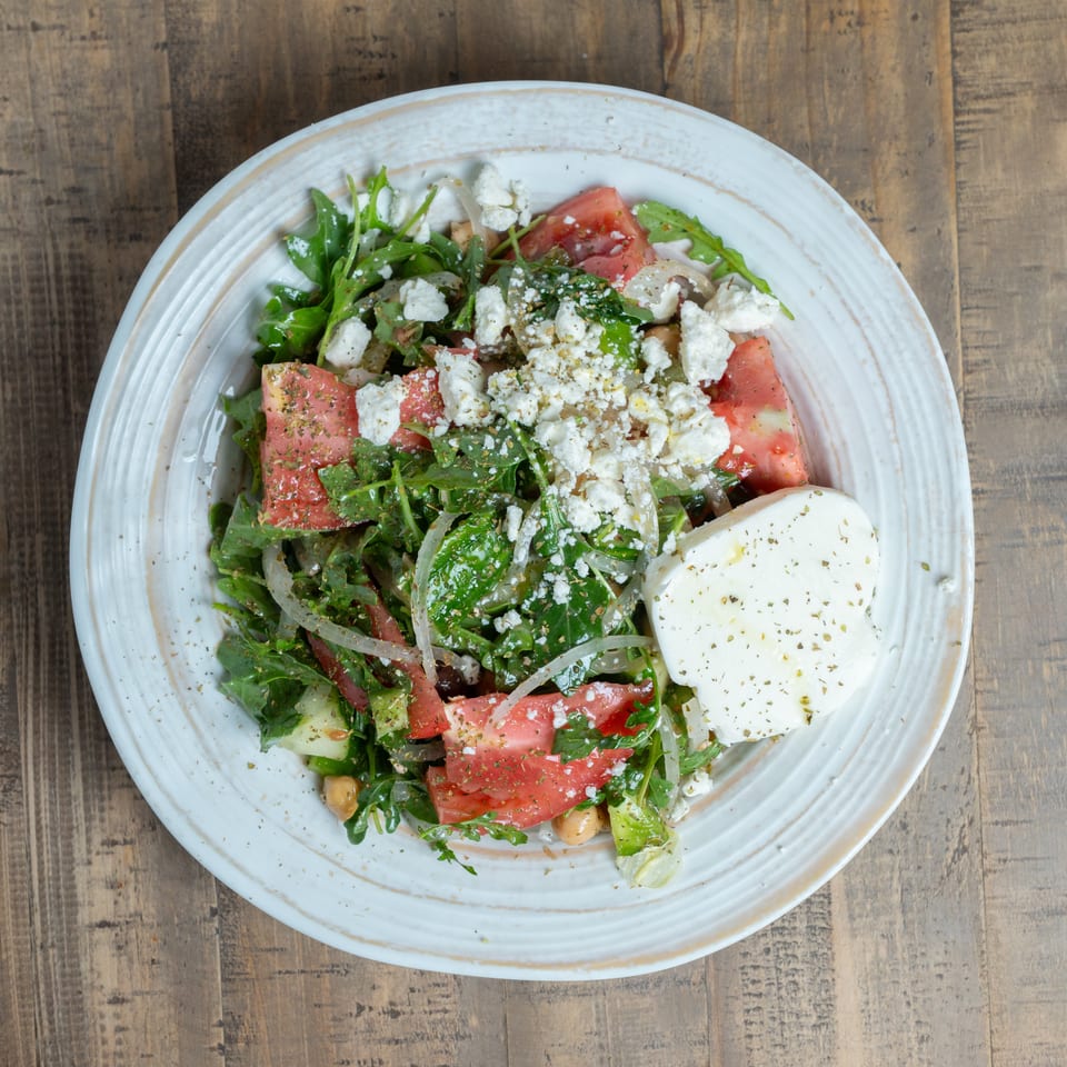 Vibrant salad with greens, tomatoes, chickpeas, crumbled feta, and a large cheese slice on a white plate.