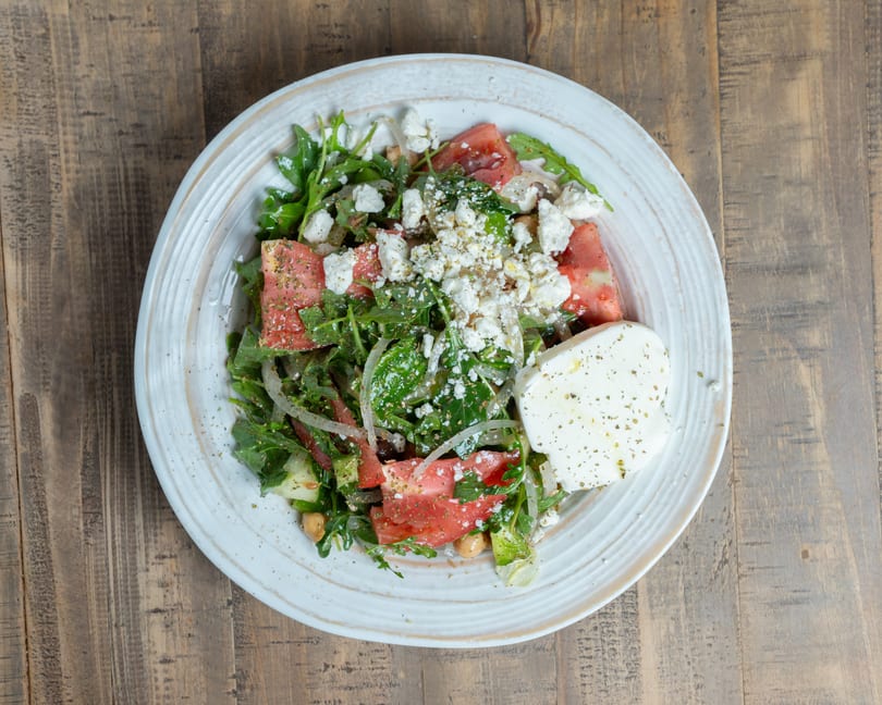 Vibrant salad with greens, tomatoes, chickpeas, crumbled feta, and a large cheese slice on a white plate.