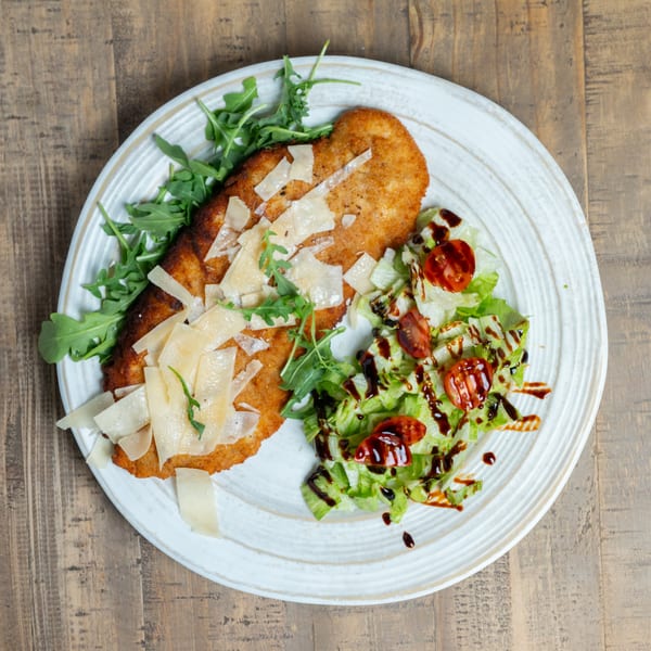 Breaded cutlet topped with Parmesan and arugula, alongside a salad with tomatoes and balsamic glaze.