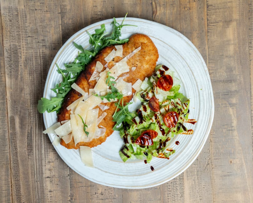 Breaded cutlet topped with Parmesan and arugula, alongside a salad with tomatoes and balsamic glaze.