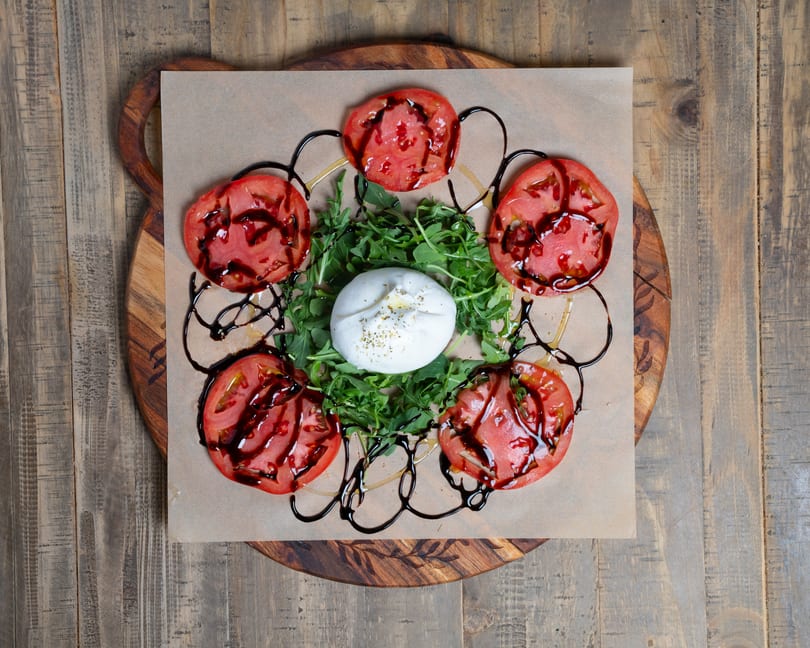 Fresh burrata, sliced tomatoes, arugula, and balsamic glaze on a rustic wooden board.