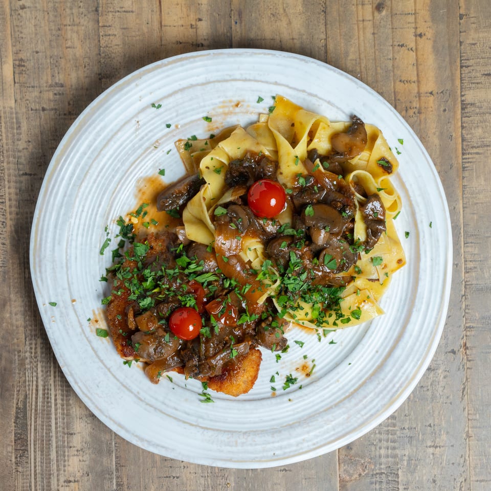Plate of pappardelle pasta with mushroom ragu, breaded cutlet, cherry tomatoes, and fresh parsley.