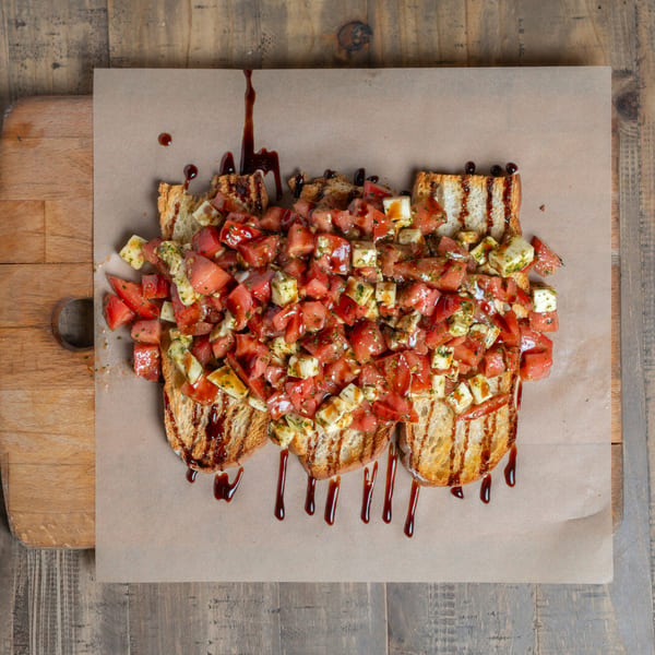 Grilled bruschetta topped with diced tomatoes, cheese, and herbs, drizzled with balsamic glaze on a wooden board.