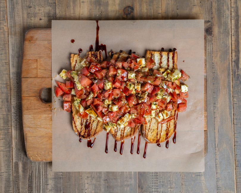 Grilled bruschetta topped with diced tomatoes, cheese, and herbs, drizzled with balsamic glaze on a wooden board.