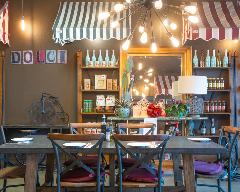Warm Italian restaurant interior with dining tables, shelves of gourmet products, and striped awnings.