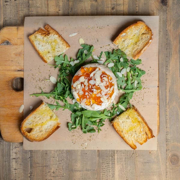 Baked brie topped with jam and almonds, surrounded by arugula and toasted bread on a wooden board.