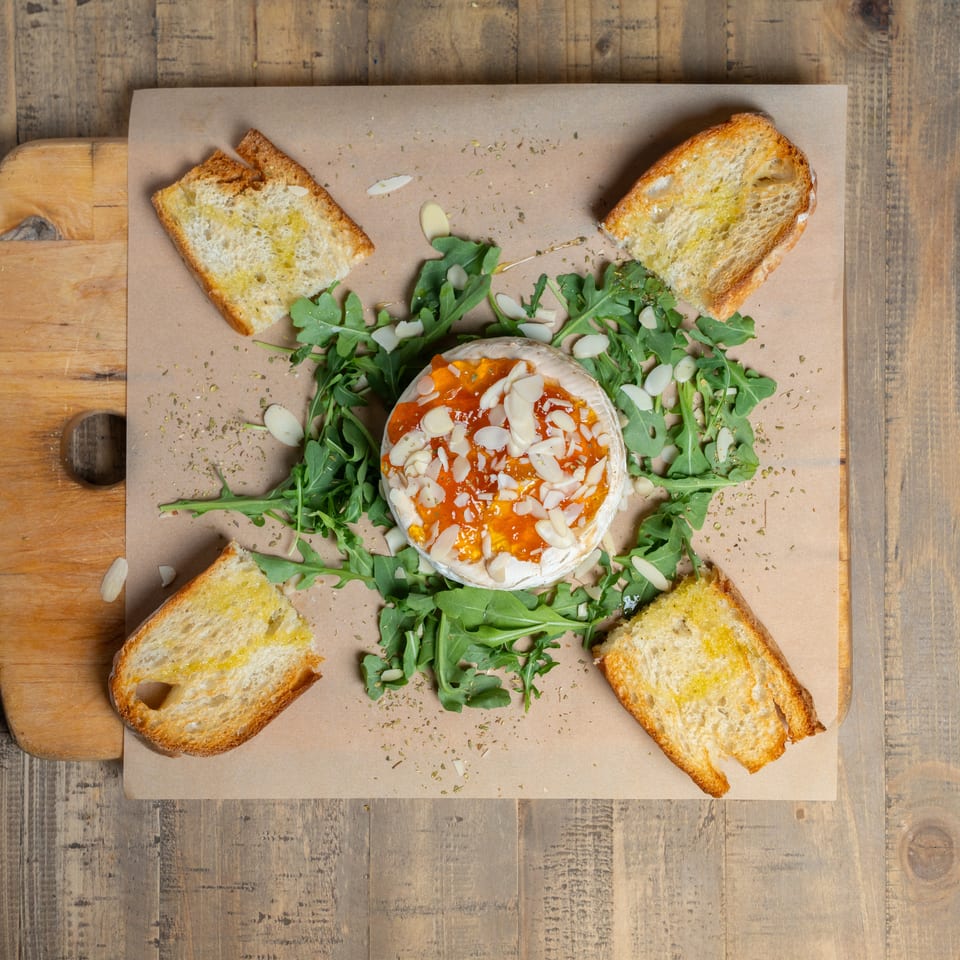 Baked brie topped with jam and almonds, surrounded by arugula and toasted bread on a wooden board.