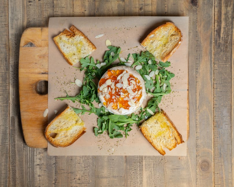 Baked brie topped with jam and almonds, surrounded by arugula and toasted bread on a wooden board.