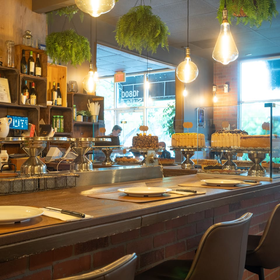 Warmly lit restaurant interior featuring a dessert display of various cakes on a brick and wood counter.