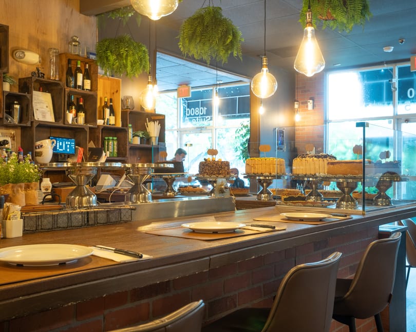 Warmly lit restaurant interior featuring a dessert display of various cakes on a brick and wood counter.